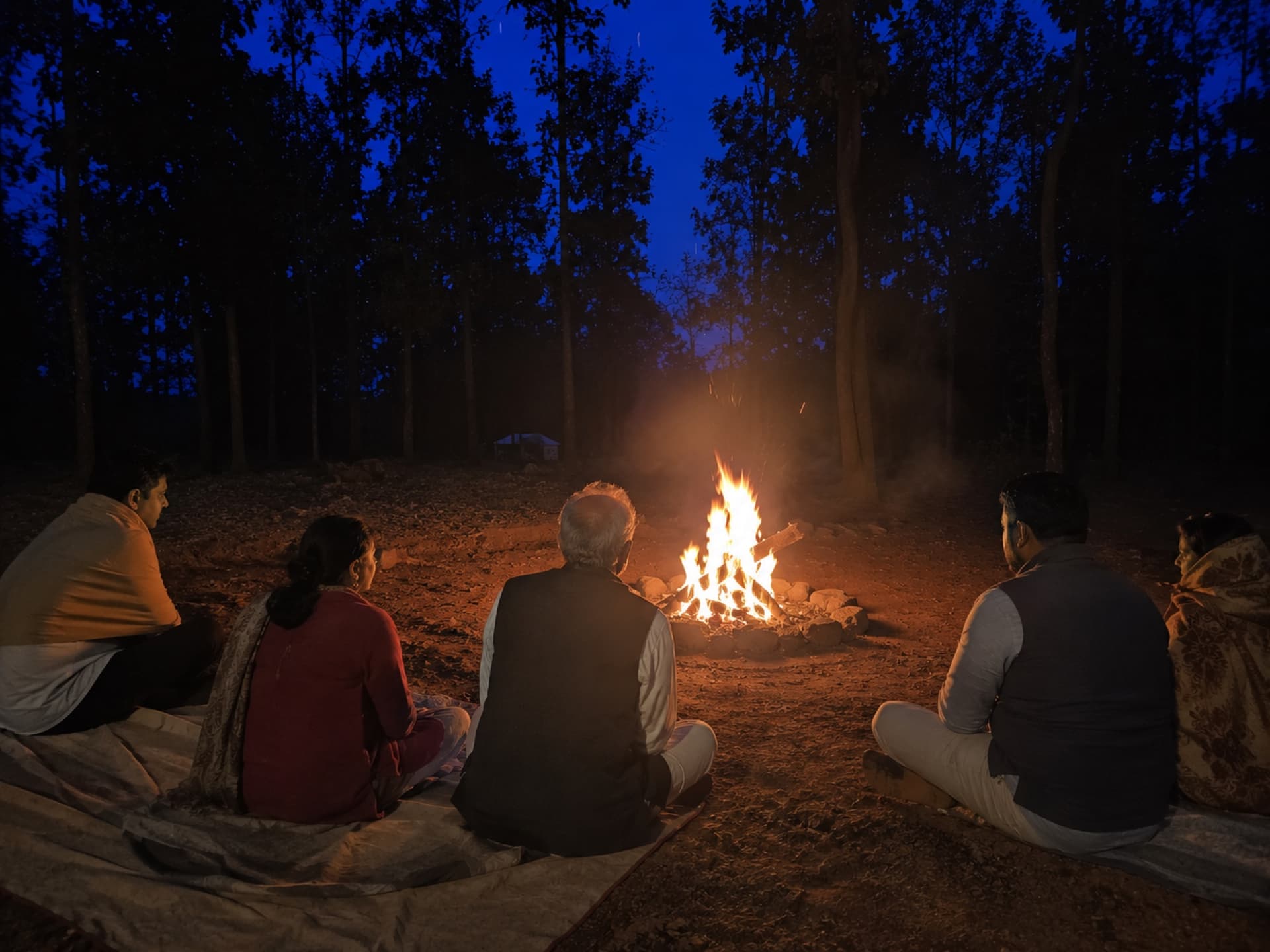Guests seated on durries around a small evening bonfire under a deep blue dusk sky