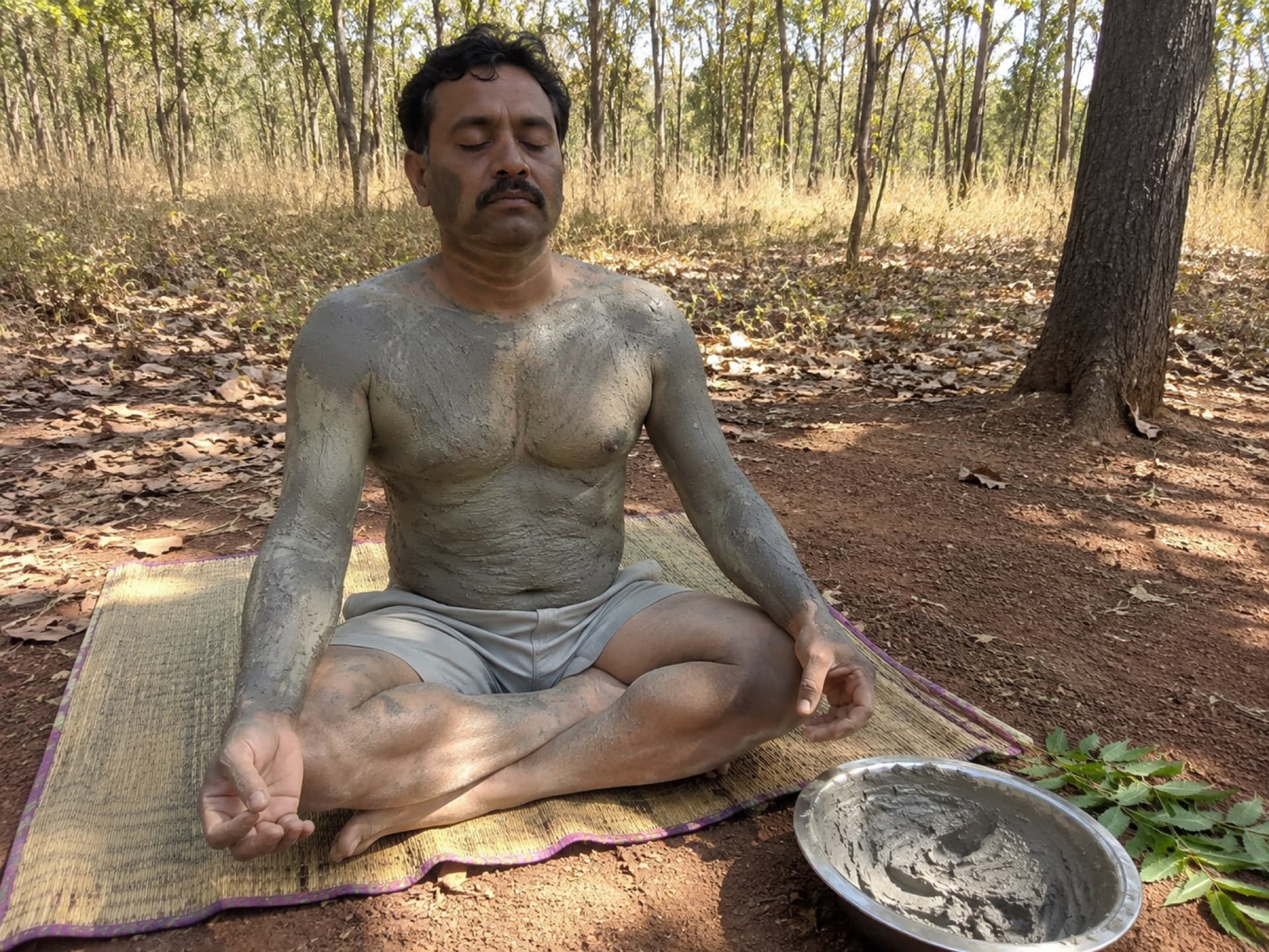 A man sits at rest under the trees, his arms and chest coated in drying river clay