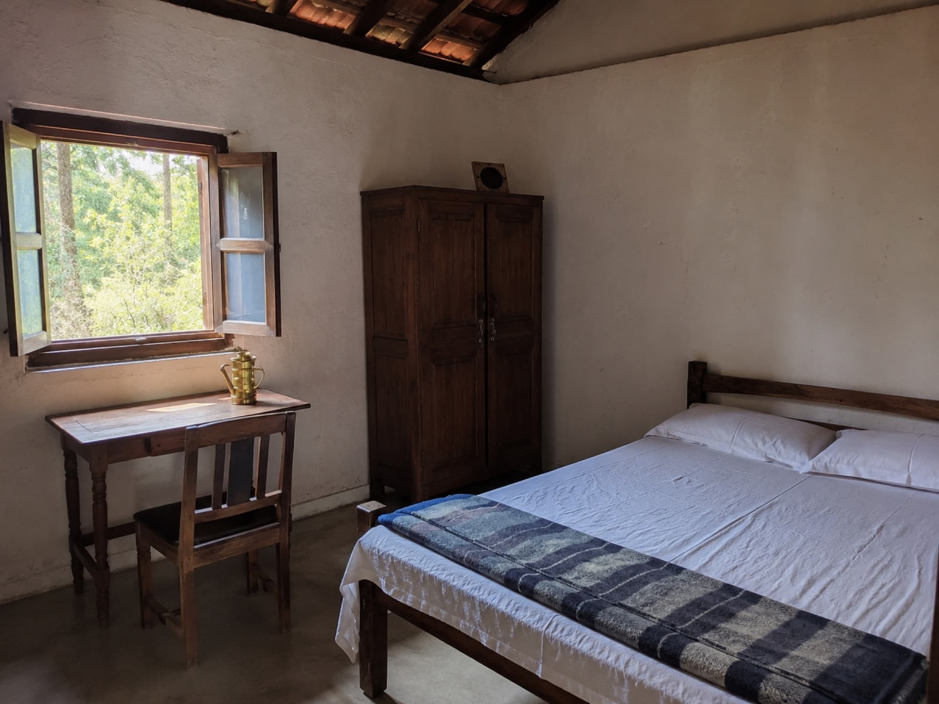 A simple cottage room — wooden bed, writing table by an open window with a forest view